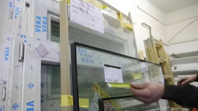 Eine Hand hält eine Isolierglasscheibe mit Etikett in der Werkstatt, daneben verpackte VEKA-Fensterprofile